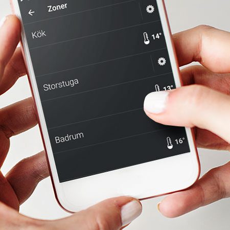 overvakning-mobil En person håller i en smartphone där en app för temperaturreglering i olika rum visas, med svenska etiketter: Kök, Storstuga och Badrum, och temperaturerna 14 °C, 13 °C och 16 °C.